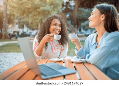 Two women sit at a table outside a cafe, enjoying their coffee and each others company. They both have cups in their hands and are smiling, engaging in conversation. A laptop rests on the table - Powered by Shutterstock