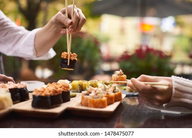 Two women sitting in asian restaurant and eating sushi set with help of chopsticks. Colleagues sharing tasty Japanese dinner with each other. Girls enjoying oriental meal from rice and seafood.