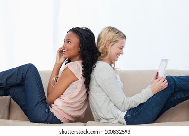 Two Women Are Sitting Back To Back On A Couch With A Tablet