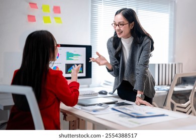 Two women are sitting at a desk with a computer monitor in front of them. One of the women is pointing at the monitor and the other woman is looking at her. Scene is one of collaboration and teamwork