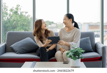 Two women are sitting on a couch, laughing together and sharing a tablet computer, in a bright living room. - Powered by Shutterstock