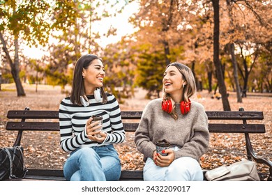 Two women sitting on a park bench with headphones and drinking coffee - Powered by Shutterstock