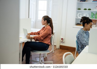 Two Women Sitting In An Open Plan Office On Teal And White Chairs Working On Computers