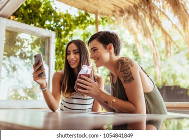 Two Women Sitting At A Restaurant Looking At Mobile Phone. Woman Showing Mobile Phone While Another Woman Enjoys A Smoothie.