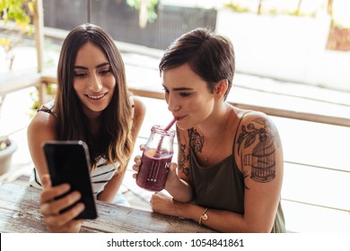 Two Women Sitting At A Restaurant Looking At A Mobile Phone. Woman Showing Mobile Phone While Another Woman Enjoys A Smoothie.