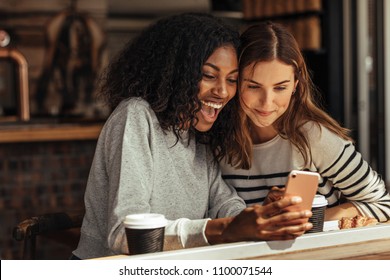 Two women sitting in a restaurant looking at mobile phone and smiling. Friends sitting at a cafe with coffee and snacks on the table looking at a mobile phone. - Powered by Shutterstock