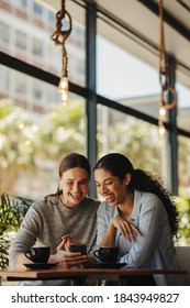 Two Women Sitting In A Restaurant Looking At Mobile Phone. Friends Sitting At A Cafe With Coffee On The Table Looking At Mobile Phone.