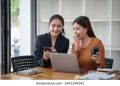 Two women are sitting at a table with a laptop and cell phones in front of them. They are smiling and laughing while looking at their devices - Powered by Shutterstock