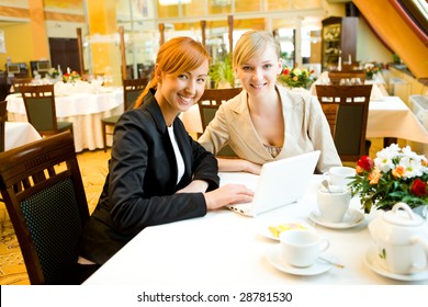 Two women sitting at table in restaurant. They're smiling and looking something on laptop.