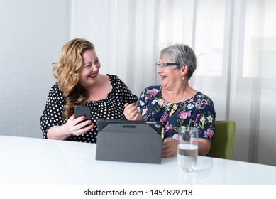 Two Women Sitting At A White Table Using Tablet And Smart Phone In Front Of White Curtains In The Background. Overweight And Mature Woman Looking At Each Other Laughing