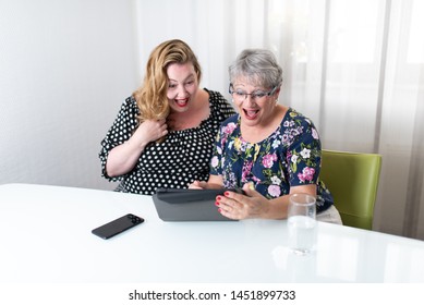 Two Women Sitting At A White Table Using Tablet And Smart Phone In Front Of White Curtains In The Background. Overweight And Mature Woman Looking Both At Screen With Open Mouth Laughing