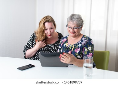 Two Women Sitting At A White Table Using Tablet And Smart Phone In Front Of White Curtains In The Background. Overweight And Mature Woman Looking At Screen Smiling 