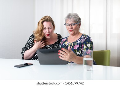 Two Women Sitting At A White Table Using Tablet And Smart Phone In Front Of White Curtains In The Background. Overweight And Mature Woman Looking Surprised At Screen 