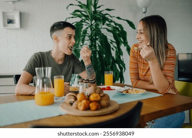 Two women are sitting at a wooden table, enjoying a healthy breakfast of scrambled eggs, pastries, and orange juice, sharing a moment of intimacy and connection in their modern apartment - Powered by Shutterstock