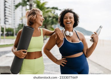 Two women in sports clothing standing on a beach promenade holding a yoga mat and a water bottle. Happy yoga friends laughing together by the ocean, enjoying a healthy lifestyle together. - Powered by Shutterstock