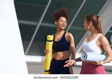 Two women in sports uniforms hold yoga mats, chatting while standing outdoors - Powered by Shutterstock