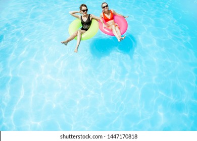 Two Women Swimming On The Inflatable Rings, Relaxing In The Water Pool Outdoors During The Summer Time, View From Above