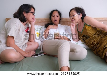 Two women tease their little sister by showing something on her sister's cell phone. both women smiled; their sister was surprised, confused.