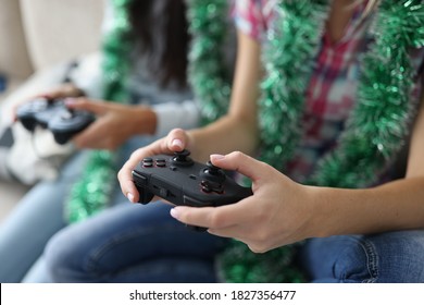 Two Women With Tinsel Around Their Necks Are Holding Joysticks Close-up. Fun Computer Games New Year And Christmas Celebration Concept.