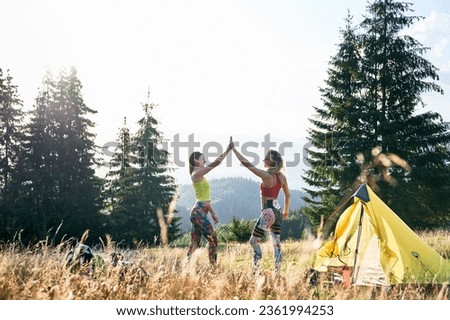 Two women tourists camping outdoors. Young women traveling in mountains, hiking. Slim, pretty females giving high five, smiling, posing in campsite near tents. Concept of beauty of nature.