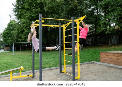 Two women are using pull-up bars in a park. One is suspended from the bar while the other climbs. The park has green grass and trees around them.