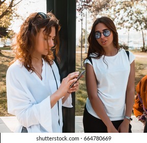 Two women waiting for a bus at a bus stop.Girls is standing on bus station.girl chatting,Girl wear stylish white shirt,attractive,pretty face,happy woman,travel mood,make up artist,tan,hipster - Powered by Shutterstock