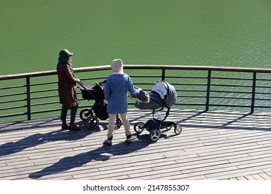 Two Women Walking With Baby Prams On Lake Embankment. Mothers Talking In City Park At Spring