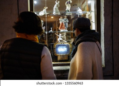 Two Women Watching Old Soviet TV In The USSR At A Shop Window With Old Porcelain Statues And Figures - Riga, Latvia