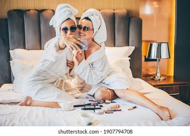Two women wearing bathrobe sitting at hotel room, making selfie and having fun a on bed in hotel room. 