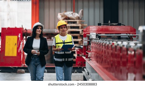 Two women wearing safety gear and holding clipboards walk through a factory. Scene is serious and focused, as the women are likely inspecting or monitoring the production process - Powered by Shutterstock