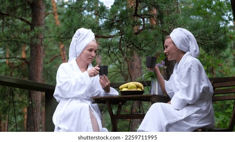 Two women in white coats sit at a wooden table on the terrace of their hotel room in the wild forest of Altai. Women drink morning coffee. Glamping in the Altai mountains on the banks of the Katun - Shutterstock ID 2259711995