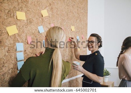 Two women are at work in an office. They are standing at a cork board and are having a discussion as they pin things up. 