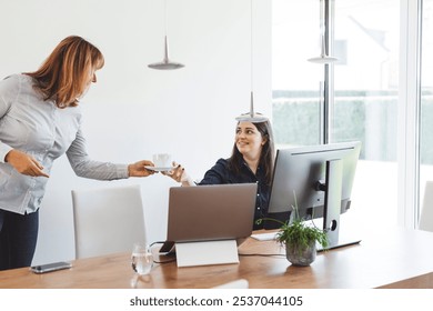 Two women working together at a modern office desk, one holding a glass of water and smiling, while the other is focused on a laptop. A cup and a small plant are on the table. - Powered by Shutterstock