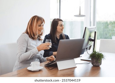 Two women working together at a modern office desk, one holding a glass of water and smiling, while the other is focused on a laptop. A cup and a small plant are on the table. - Powered by Shutterstock