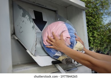 Two Women's Hands Putting A Plastic Bag Full Of Used Clothing Into An Old White Clothes Donation Bin Or Container For Shoes And Textiles.Concept Donation And Reused Recycle