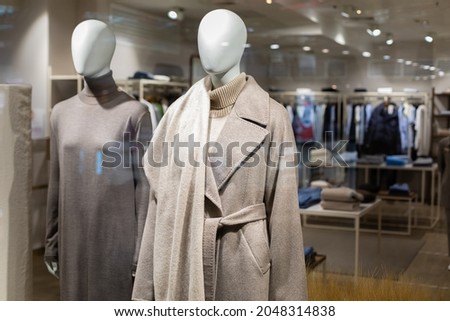 Two women's mannequins in the window of a large outerwear store. Autumn winter coat made of wool of natural color with a belt and winter long knitted dress. Shop hall in the background