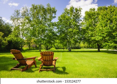 Zwei hölzerne Beistellstühle auf üppigem, grünem Rasen mit Bäumen