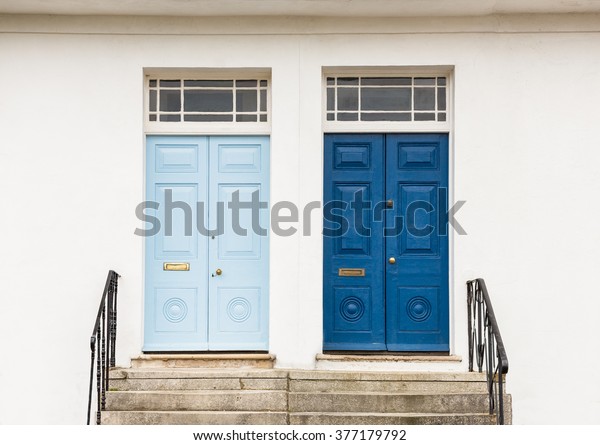 Two wooden blue, one
dark blue, one light blue, Georgian style font doors to homes,
against a white wall at the top of steps with metal banisters and
windows at the top.