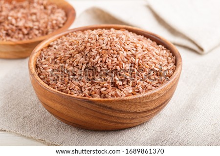 Two wooden bowls with unpolished brown rice on a white wooden background and linen textile. Side view, close up, selective focus.