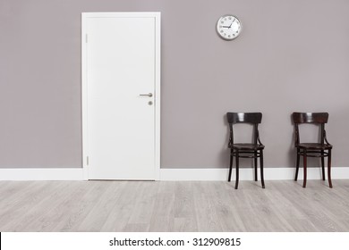 Two Wooden Chairs In A Waiting Room With A Clock Hanging On The Wall Above Them