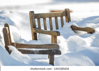 Two wooden frame chairs buried in snow on a sunny winter cold day. Several feet of snow completely cover the chairs with only the back and armrest showing out of the white fluffy snow - Powered by Shutterstock