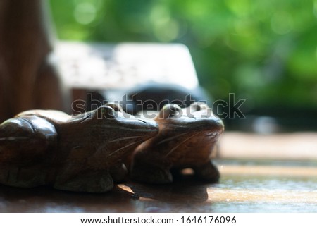 Two wooden frogs places together on the table with sunlight shining to them from the glass window, its background is blurred green plant