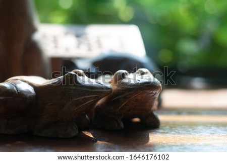 Two wooden frogs places together on the table with sunlight shining to them from the glass window, its background is blurred green plant