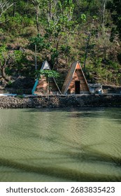 Two wooden triangular huts stand beside the water, blending rustic architecture with peaceful nature in a riverside setting. 