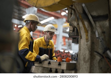Two workers are busy operating a machine in a factory. They wear helmets and gloves as they handle the equipment. The factory shows an industrial setting with bright lights.