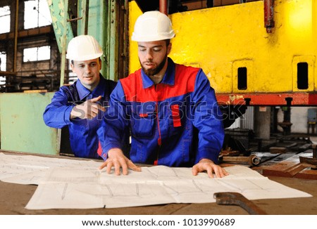 Two workers in construction helmets discuss a plan, blueprint or industrial project in the background of the plant