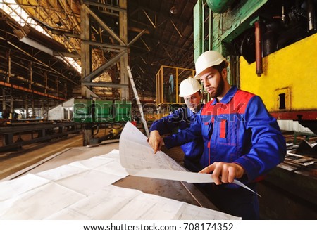 Two workers in construction helmets discuss a plan, blueprint or industrial project in the background of the plant