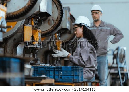 Two workers are in a factory setting. One is seated and using machines while the other stands nearby. They wear helmets and protective clothing. The area is filled with various equipment.
