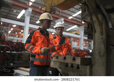 Two workers are focused on a machine in a factory. They wear safety helmets and bright jackets. The setting shows industrial equipment and a busy workspace.