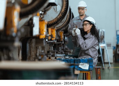 Two workers are focused on using machines in a factory. One worker checks parts while another stands by. The environment is busy with tools and equipment around them.